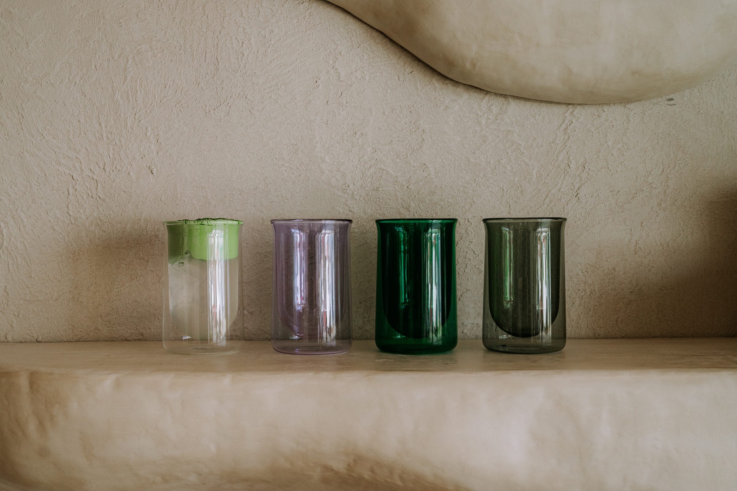 Four FLUR double-walled glasses in green, purple, emerald and smoke on a plaster ledge